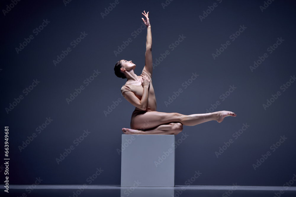 Fototapeta premium Graceful. tender, attractive woman, ballerina in beige bodysuit posing, sitting on cube against blue studio background. Concept of classical dance, art and grace, beauty, choreography, inspiration