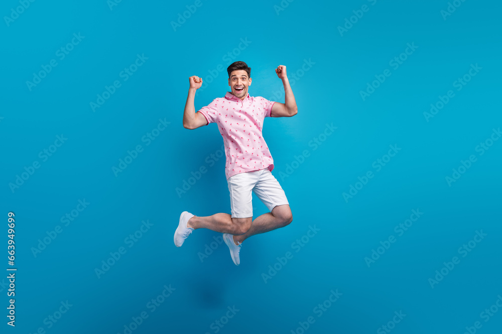 Full body portrait of crazy energetic young man jumping raise fists accomplishment isolated on blue color background Full body portrait of crazy energetic young man jumping raise fists accomplishment isolated on blue color background