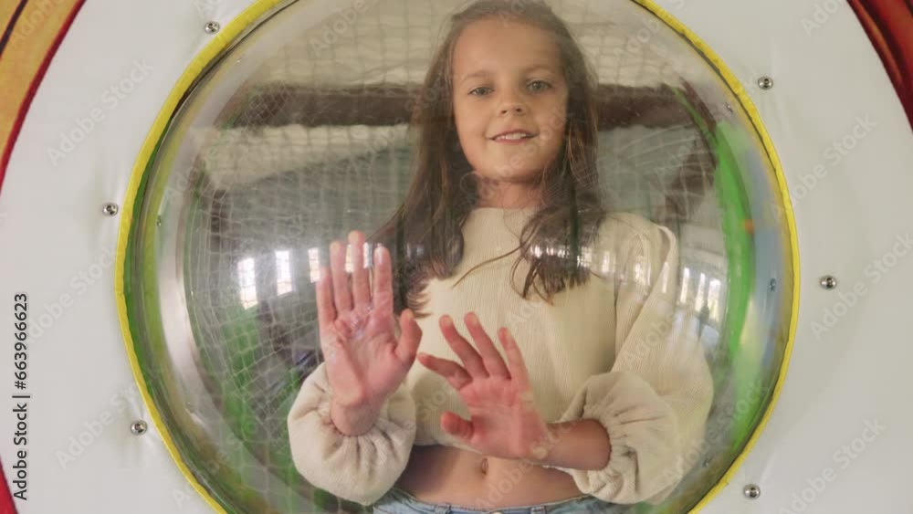 Brown haired girl playing in playground indoor looking from window in round tunnel in children's playroom expressing happiness. Brown haired girl playing in playground indoor looking from window in round tunnel in children's playroom expressing happiness.
