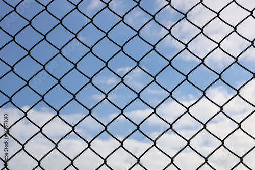 Blue sky behind the metal mesh fence. Wire mesh on background of blue sky and white clouds. Rusty wired fence, metallic net.