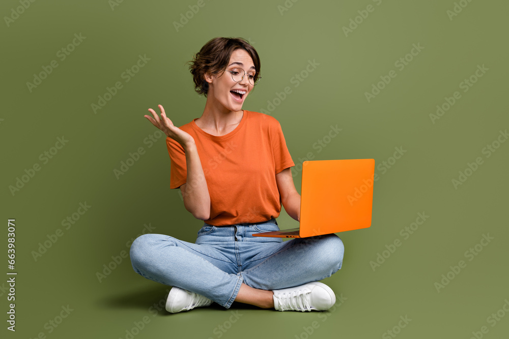 Full length photo of nice girl dressed t-shirt in eyewear sit on floor talk on laptop video conference isolated on green color background Full length photo of nice girl dressed t-shirt in eyewear sit on floor talk on laptop video conference isolated on green color background