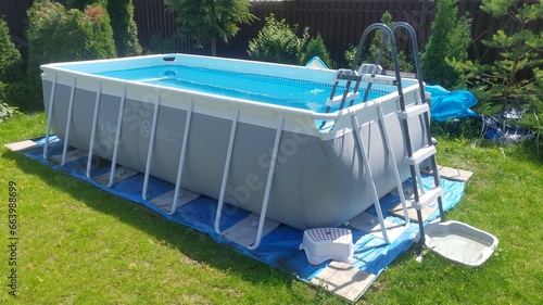 The gray rectangular frame swimming pool stands on a grassy lawn. The water is cleaned with a filter. Next to the metal fence there are thuja trees. The pool is made of PVC. Sunny summer weather photo