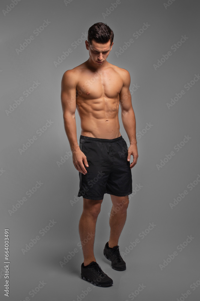 Full length image of a man with an athletic and fit body, posing in the studio with a bare torso, showing six abs pack,  on the grey background.