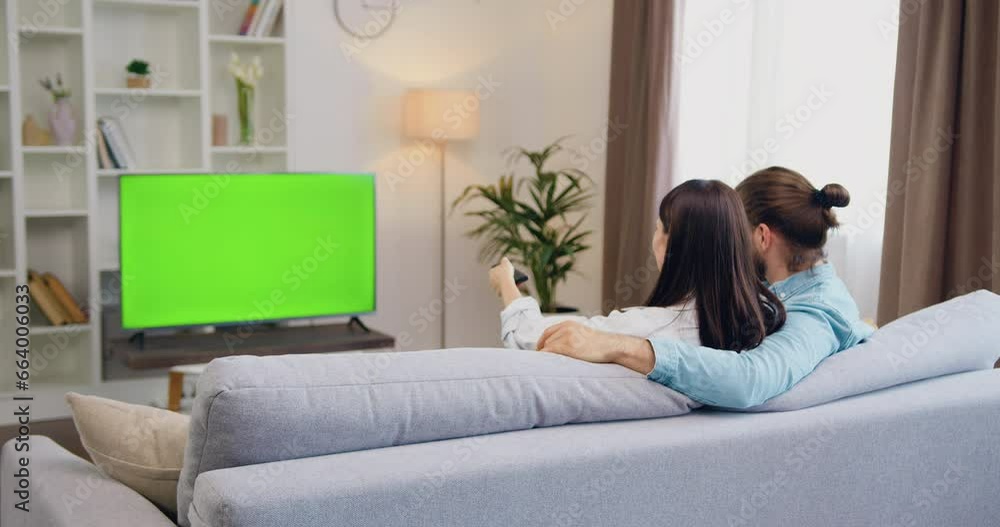 Caucasian happy couple man and woman having fun, sitting on sofa in living room watching at TV monitor with green chroma key screen. Woman switching channels with remote control. Resting at home in Caucasian happy couple man and woman having fun, sitting on sofa in living room watching at TV monitor with green chroma key screen. Woman switching channels with remote control. Resting at home in
