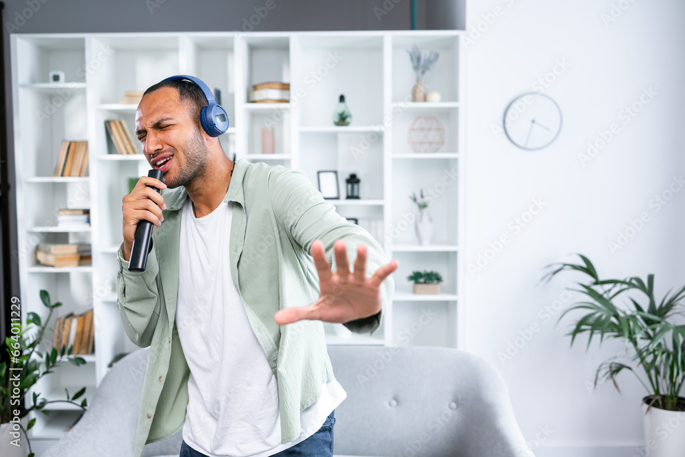 Carefree Singer. Cheerful young guy singing favorite song using remote control as microphone, dancing wearing wireless headset. Excited man having fun at home