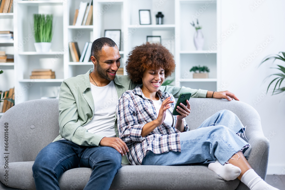 Overjoyed young couple sit rest on couch at home have fun watching video on smartphone together. Happy man and woman enjoy weekend in cozy living room - obrazy, fototapety, plakaty 