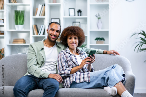 Smiling husband and wife sit on couch using smartphone, happy young couple relax on sofa look at camera