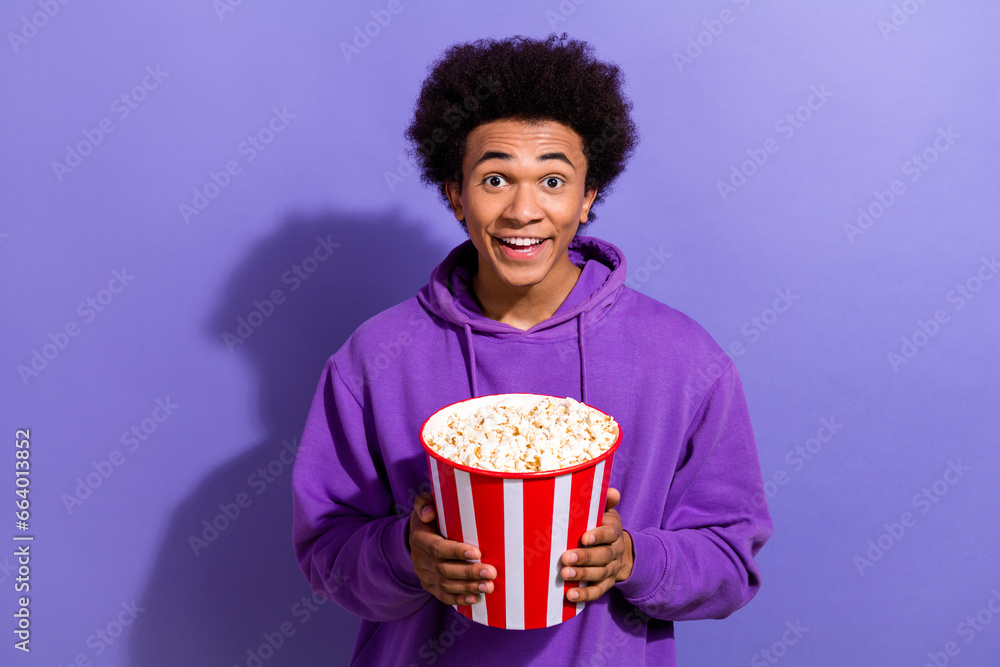 Photo of astonished positive person toothy smile hands hold big popcorn bucket isolated on violet color background Photo of astonished positive person toothy smile hands hold big popcorn bucket isolated on violet color background