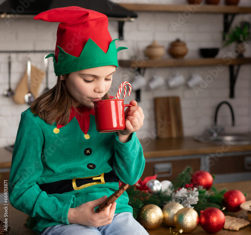 Little girl in elf costume holding chocolate biscuit and mug with candy cane in it in hand. Child drinking beverage, sitting on kitchen counter decorated with Christmas ornaments, gold and red baubles