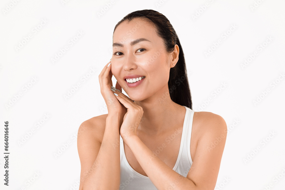 Smiling asian middle aged woman touching face, black ponytail, white backdrop Smiling asian middle aged woman touching face, black ponytail, white backdrop
