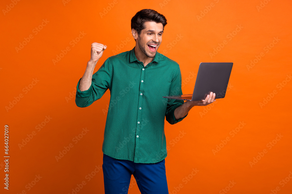 Photo of awesome young guy wear trendy green shirt hold laptop celebrate his promotion career success isolated on orange color background Photo of awesome young guy wear trendy green shirt hold laptop celebrate his promotion career success isolated on orange color background