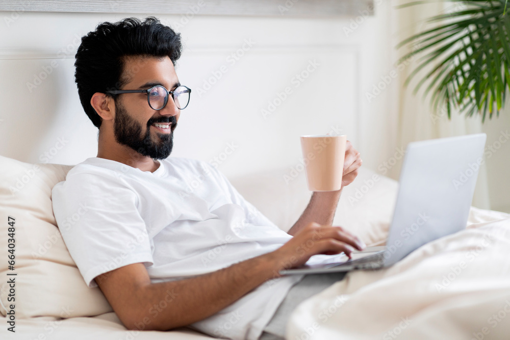 Obraz premium Young Indian Man Using Laptop And Drinking Coffee While Sitting In Bed