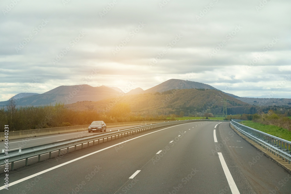 A lonely car drives along a motorway in France in the rays of the setting sun against the backdrop of mountains.