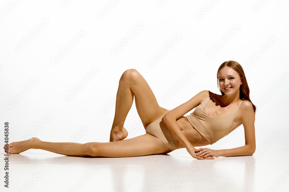 Elegant, beautiful young girl with fit, slim body posing in floor in beige underwear against white studio background. Concept of natural beauty, body and skin care, health, sport, wellness. Elegant, beautiful young girl with fit, slim body posing in floor in beige underwear against white studio background. Concept of natural beauty, body and skin care, health, sport, wellness.