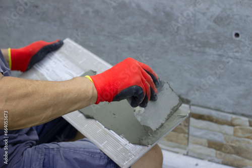 A male builder applies adhesive tile to lay it on the enclosing wall of the building from the outside. A construction contractor lays facing tiles on the fence of a private house.