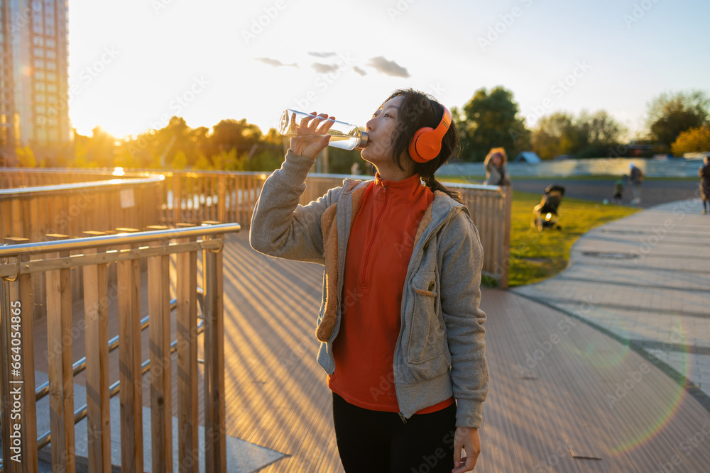 young beautiful and happy Asian Korean woman in sportswear listening to music with headphones and holding bottle of water after workout on city bridge in healthy lifestyle concept