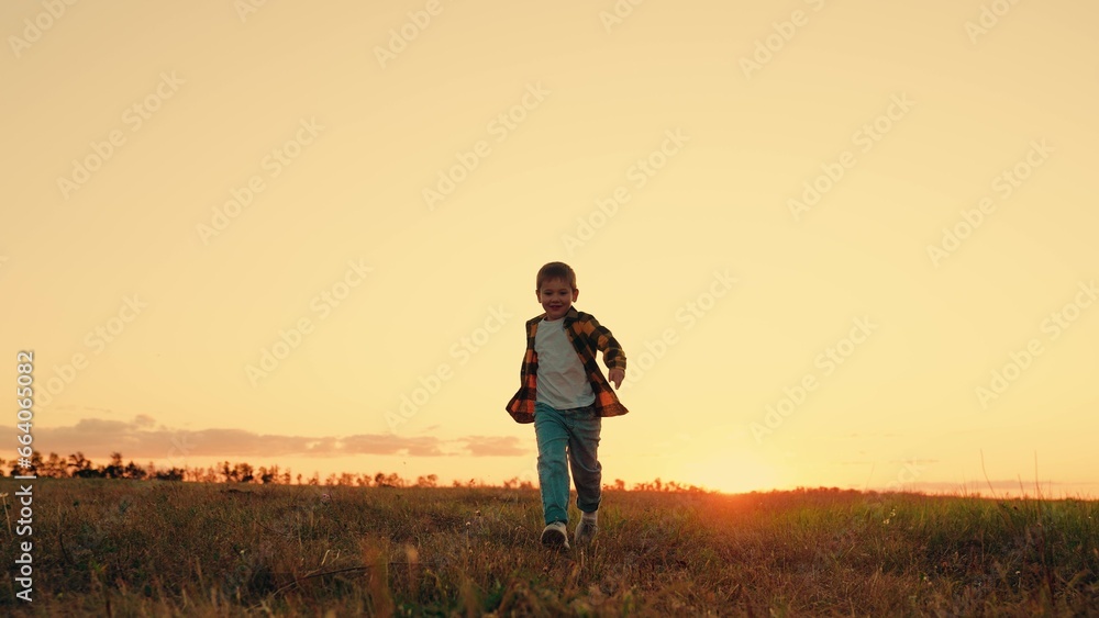 Joyful little boy running at sunset. Kid is running across field. Child boy runs through green grass in sun. Childhood dream happiness concept. Happy child playing in nature. Happy family. Dream kid