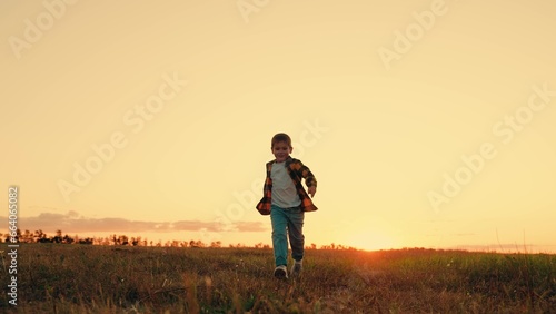 Joyful little boy running at sunset. Kid is running across field. Child boy runs through green grass in sun. Childhood dream happiness concept. Happy child playing in nature. Happy family. Dream kid
