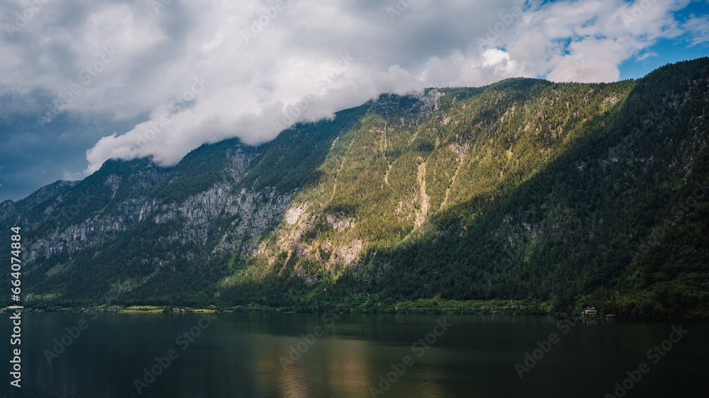 A mountain on the shore of the alpine lake Hallstätter See illuminated by the afternoon sun shining through the rain clouds. A house on the edge of the lake.