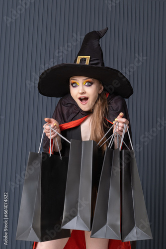 Surprised young woman in witch hat with black shopping bags. Seasonal discounts. Halloween sale. Dark background. Black friday