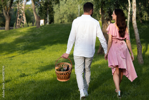 Concept of celebration happy valentines day outdoors. Rear view couple dressed stylish elegant clothing walking for a picnic. Young family bring wicker basket to the lunch in park