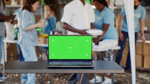 Digital laptop with blank green screen display is placed on table while volunteers help the less fortunate. Wireless computer in the foreground with isolated chromakey mockup template. Tripod shot.