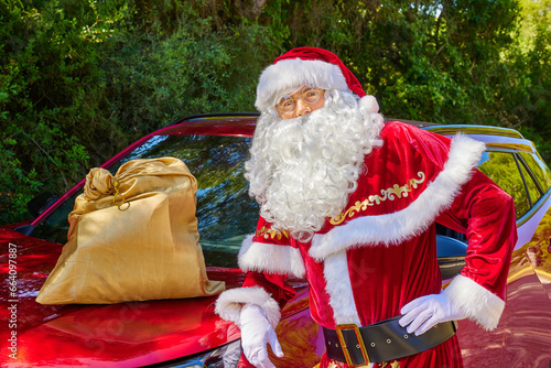 Gorgeous Santa Claus in glasses stands next to a red car in a green forest with the bag of gifts. Close up © Anna Baranova