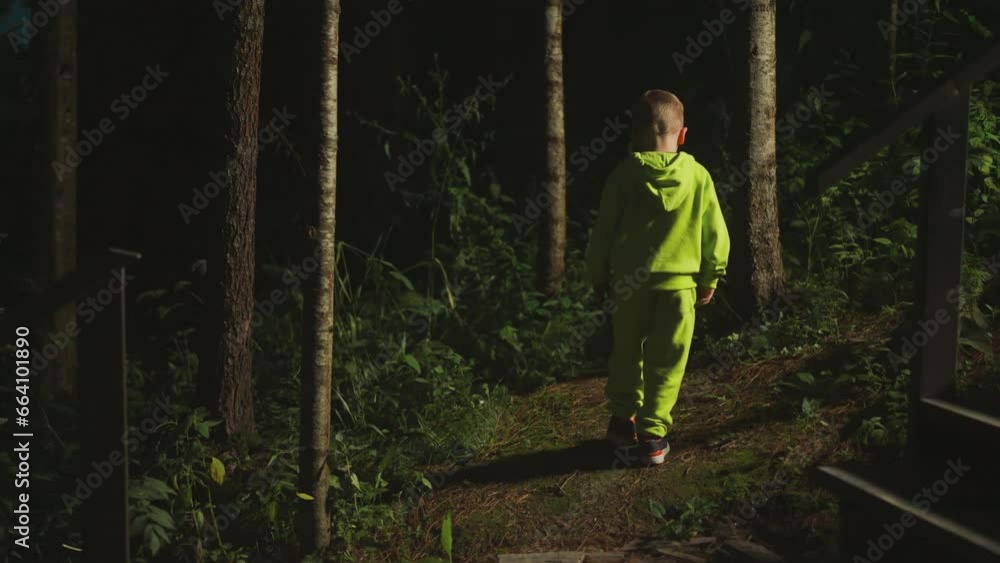 Curious little boy in bright tracksuit goes alone into forest. Child goes into evening thickets from stairs of home in search of adventure. Nature environment Curious little boy in bright tracksuit goes alone into forest. Child goes into evening thickets from stairs of home in search of adventure. Nature environment