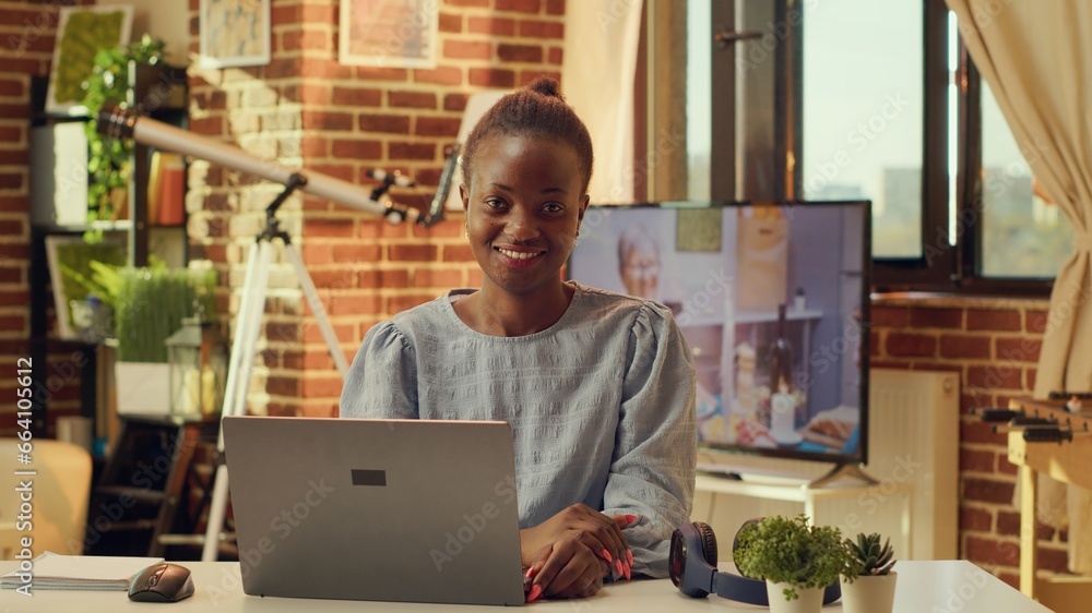 Fototapeta premium Portrait of freelancer in warm lit room working on online project tasks at desk, solving activities remotely at home. African american woman with blogger job, e business freelance work.
