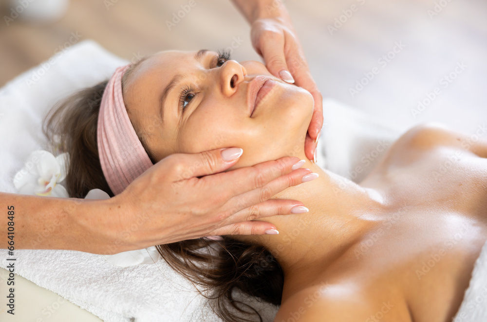 Female masseuse giving young woman client rejuvenating facial massage