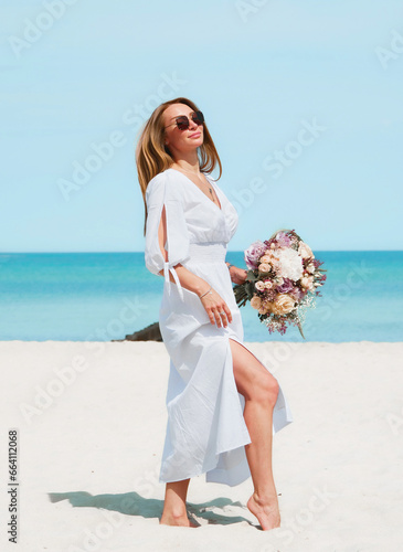 Obraz na płótnie Joyful Young Woman Holding a Bouquet of Flowers on a Beach with Blue Sky Background w salonie