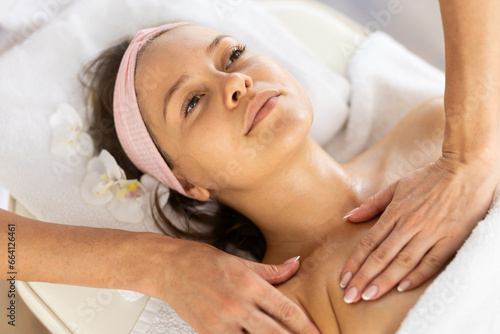 Girl enjoys and rests during decollete and neck massage procedure in beauty salon  professional woman performs relaxing stroking and pinching while working out problem areas of low neck.