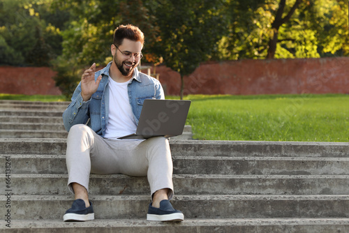 Handsome young man with laptop sitting on concrete stairs outdoors. Space for text
