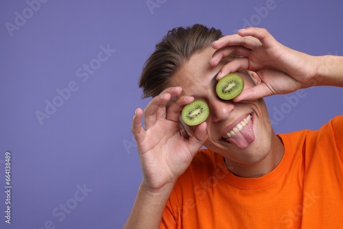 Smiling man covering eyes with halves of kiwi and showing tongue on violet background, closeup. Space for text