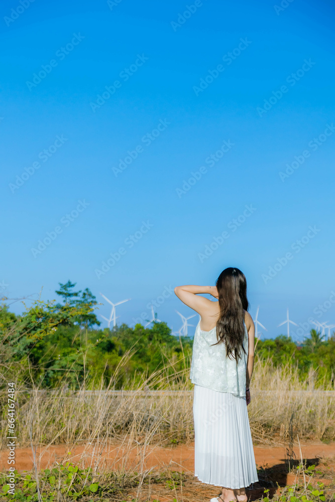A Girl stands against the wind turbine generate electricity, fan fields on the green grass over the blue clouded sky in Ea H'leo district, Dak Lak province, Vietnam. A Girl stands against the wind turbine generate electricity, fan fields on the green grass over the blue clouded sky in Ea H'leo district, Dak Lak province, Vietnam.