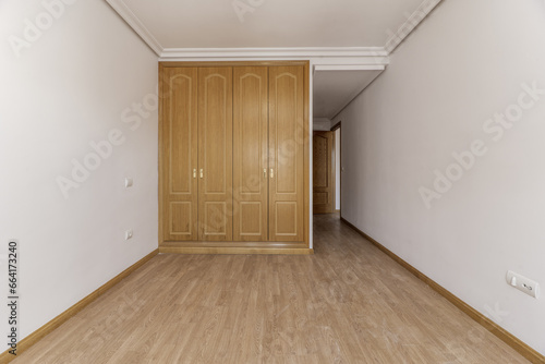 Empty bedroom with a built-in three-door oak wardrobe with gold handles  skirting boards of the same material  plain white painted walls and light wooden floor