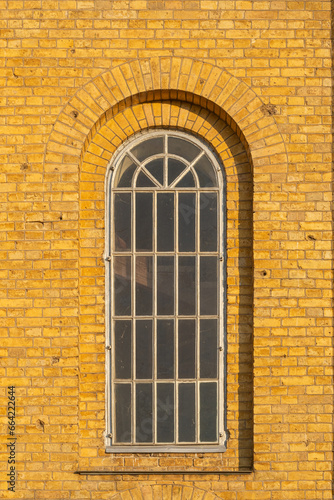 A large wooden arched white window on an old yellow brick wall of a 19th century church facade.