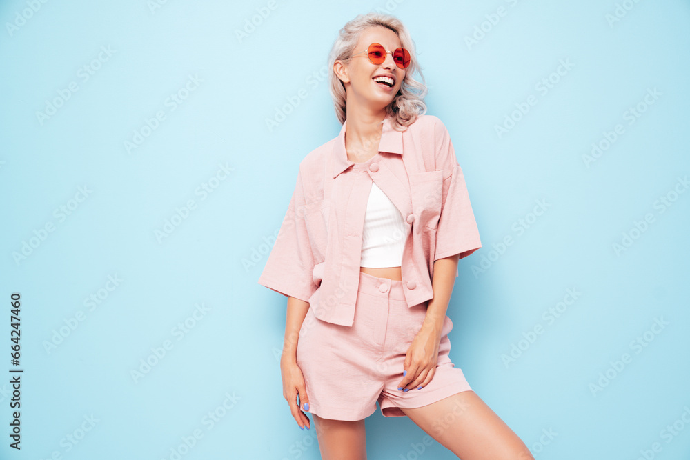 Young beautiful smiling female in trendy summer pink shorts and t-shirt. Carefree woman posing near blue wall in studio. Positive blond model having fun. Cheerful and happy Young beautiful smiling female in trendy summer pink shorts and t-shirt. Carefree woman posing near blue wall in studio. Positive blond model having fun. Cheerful and happy