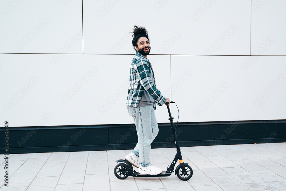 Trendy smiling bearded man in casual clothes riding electric scooter in urban background. Handsome model posing in the street near wall. Hipster guy with curly hairstyle