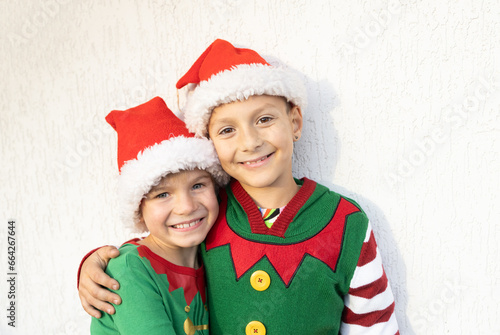 two smiling cute boys in Santa hats and elf costumes hug each other on a light background. Children rejoice on Christmas Eve  expecting gifts and miracles. the winter vacation. Family weekend