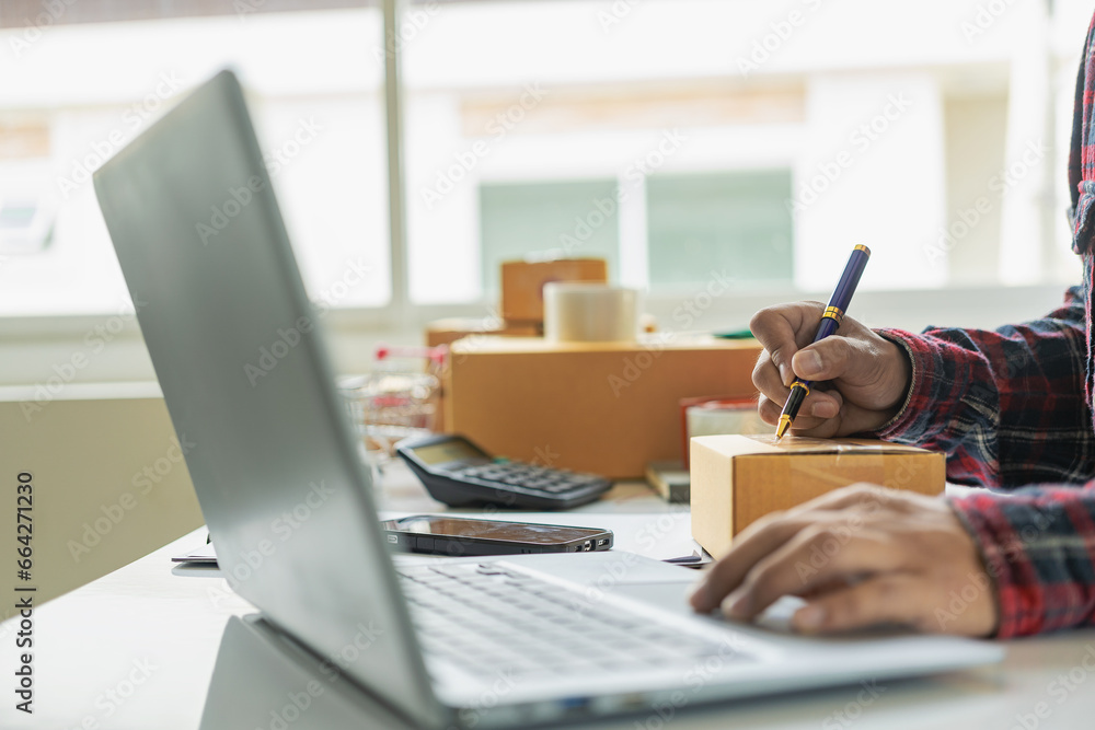 Fototapeta premium Small business startup SME business owners use smartphones or tablets to receive and monitor online orders to prepare product boxes for delivery. Close-up pictures
