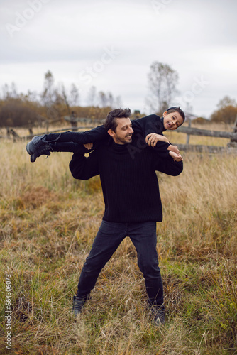 portrait stylish father and son in black clothes holding his child on his shoulders in a field with an old wooden horse fence in the fall. funny family