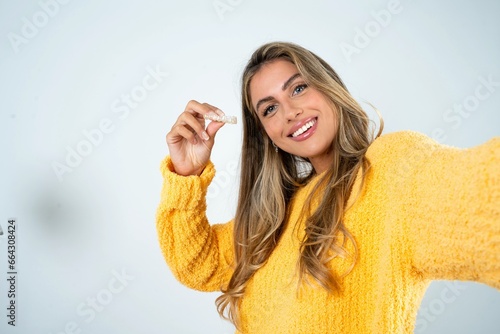 Beautiful caucasian woman wearing yellow jumper make selfie holding an invisible braces aligner, recommending this new treatment. Dental healthcare concept.