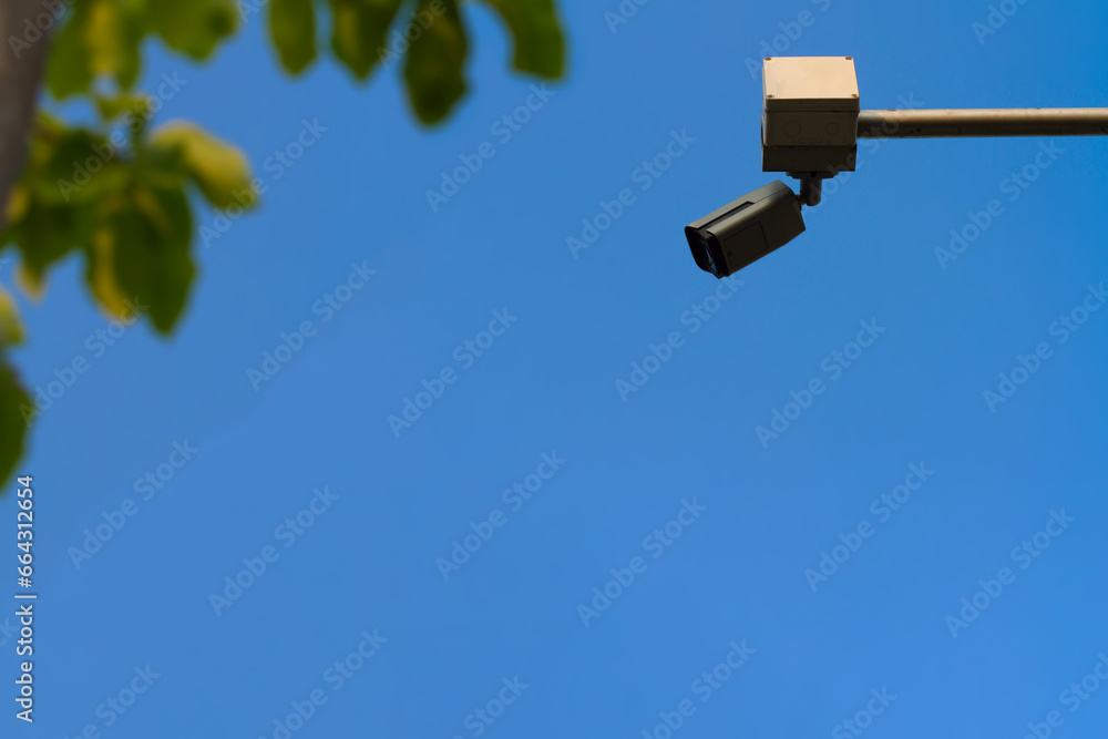 Low angle view of the surveillance security camera on steel pole in outdoor public car park with clear blue sky and blurry green leaves tree on foreground. Low angle view of the surveillance security camera on steel pole in outdoor public car park with clear blue sky and blurry green leaves tree on foreground.
