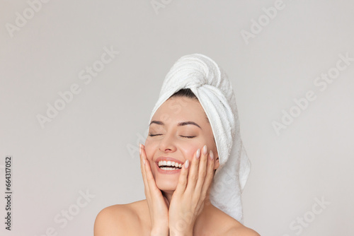 a young pretty Caucasian woman with a white towel on her head laughs, closes her eyes and gently touches her face with her hands. Isolated on a light background.