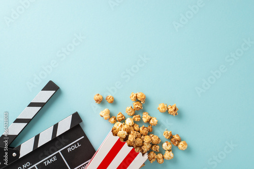 Film debut concept. Top view of moviemaker's clapperboard, and tasty striped popcorn container on soft blue backdrop with room for your message or advertisement