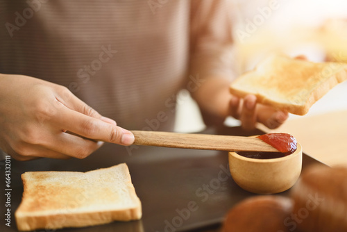 Cropped shot of woman spreading strawberry jam on slice of bread while preparing breakfast in kitchen