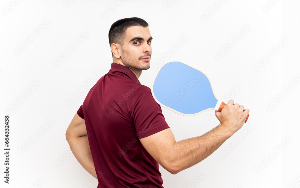 A handsome young man poses in sportswear on a white studio background with a blue pickleball racket. The man looks at the camera while resting the racket on his shoulder. Pickleball concept. A handsome young man poses in sportswear on a white studio background with a blue pickleball racket. The man looks at the camera while resting the racket on his shoulder. Pickleball concept.
