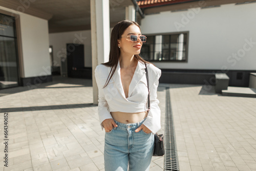 Cool fashionable American prett woman model with vintage sunglasses in trendy outfit with jacket and blue jeans with bad walks on the street on sunny day. Beautiful girl photo