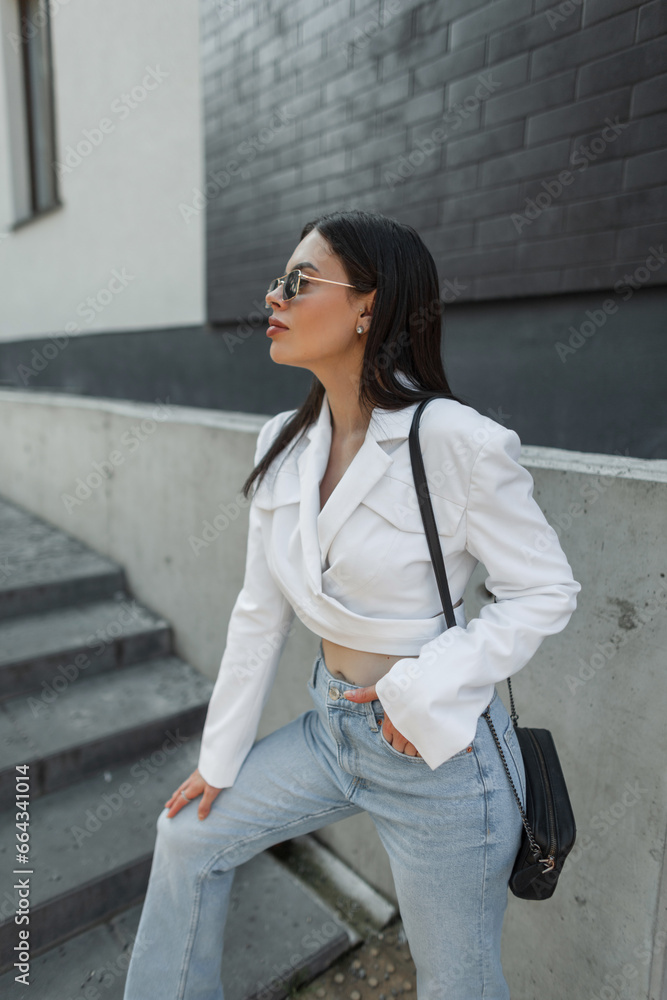 Fashionable beautiful young stylish girl with sunglasses in fashion casual clothes with a white blazer and vintage blue jeans with a bag in the city near a brick black wall Fashionable beautiful young stylish girl with sunglasses in fashion casual clothes with a white blazer and vintage blue jeans with a bag in the city near a brick black wall